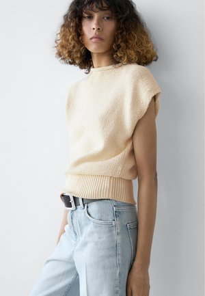 Vrouw met krullend haar die een mouwloos beige gebreid topje en lichtblauwe spijkerbroek draagt, staand tegen een effen witte muur.