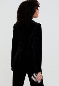 Schwarzer Samtblazer mit taillierter Silhouette, Revers mit Kerbe und Knopfdetail. Das Model hält eine gestreifte, verzierte Clutch.