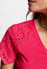 Camiseta fucsia con mangas de encaje bordadas, con patrones recortados y un cuerpo de algodón suave. Escote redondo y forma ajustada.