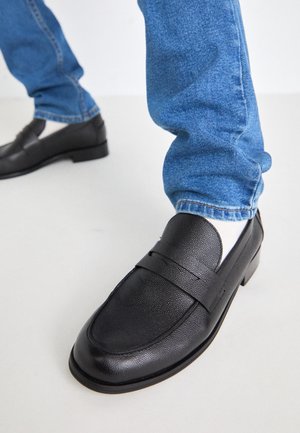 Mocassins en cuir noir avec une surface texturée, un talon plat et une accent de sangle unique. Assortis à un jean en denim bleu.