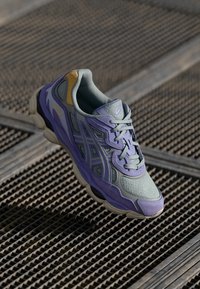 Paarse en grijze hardloopschoen met gaas en synthetische overlays, voorzien van een gevoerde kraag en vetersysteem, en een gestructureerde rubberen zool.