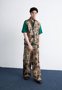 Καμουφλάζ γιλέκο και ταιριαστά cargo παντελόνια σε καφέ και πράσινα μοτίβα; με στρώση από πράσινη μπλούζα; μαύρα παπούτσια με κόκκινες λεπτομέρειες.