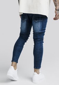Jeans ajustados en denim azul oscuro, con un lavado desgastado, diseño de cinco bolsillos y detalles de costura, combinados con zapatillas blancas.