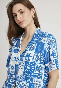 Chemise à manches courtes bleu et blanc mettant en avant un collage de motifs marins, incluant des coquillages, des vagues et des coraux, sur un tissu léger.