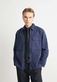 Tumed sinise värviga tõmblukuga jakk kahe esipocketiga, valmistatud pehmest kangast. Kandmiseks musta särgi ja lipsu peal, rinnal on diskreetne logo.