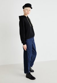 Sudadera negra con cordones dorados, combinada con unos vaqueros de mezclilla azul holgados. Llevada con botas de tobillo negras con cordones y una gorra negra.