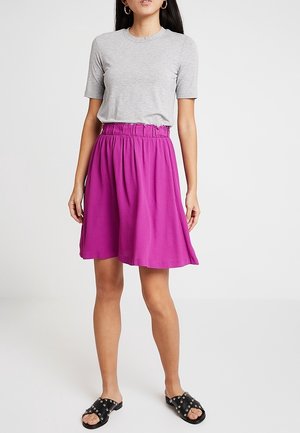 Lila, knielanger Rock mit einem elastischen Bund, kombiniert mit einem enganliegenden grauen T-Shirt. Schwarze Rutsch-Sandalen mit Nieten runden das Outfit ab.