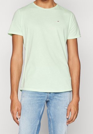 Mężczyzna w jasnozielonej koszulce z krótkim rękawem i jasnoniebieskich, postrzępionych dżinsach, stojący na tle jednolitego jasnego tła.