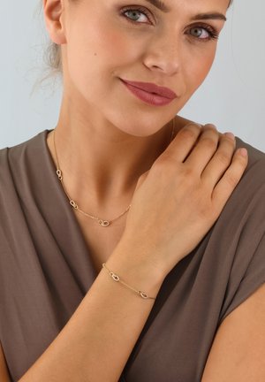 Goldfarbene Halskette und Armband-Set mit ineinander greifenden ovalen Gliedern; feines Kettendesign. Auf einem neutralen Stoffhintergrund getragen.