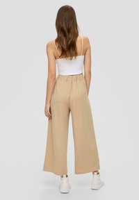 Beige wijde broek met een elastische tailleband, gecombineerd met een witte aansluitende tanktop. Het model draagt witte platform sneakers.