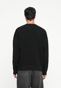 Svart ribbestrikket genser med lange ermer og rund hals, med teksturert stoff og en avslappet passform. Brukt sammen med mørkegrå jeans.