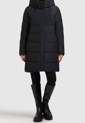 Czarny długi pikowany płaszcz z kapturem i sznurkami, noszony z czarnymi legginsami i czarnymi botkami za kolano na masywnej podeszwie, na neutralnym tle.