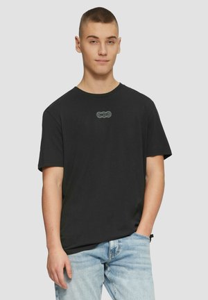 Czarny bawełniany t-shirt z okrągłym dekoltem. Zawiera małą szaro-skalową grafikę na piersi. Noszony z jasnoniebieskimi dżinsami.