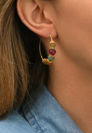 Boucle d'oreille en or avec des perles vertes, violettes et brunes portée sur l'oreille d'une femme, avec des cheveux châtain clair visibles et un col en denim bleu.