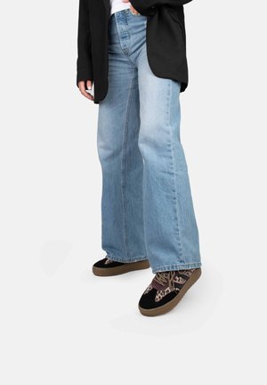Jeans évasés bleu clair avec une finition délavée, associés à des chaussures noires à motifs avec des semelles et des lacets marron. Veste noire en dessus.