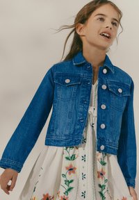 Chaqueta de denim azul con mangas largas, bolsillos frontales y botones plateados. Lleva puesta sobre un vestido blanco con bordados florales coloridos.