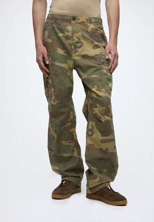 Homme portant un pantalon cargo camouflage ample et des baskets marron, debout, les mains détendues le long du corps.