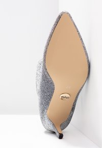 Pompe à talons aiguilles argentée scintillante avec un bout pointu, un dessus texturé et une semelle beige lisse présentant un détail de logo doré. Taille 37.