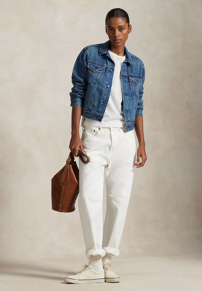 Donna che indossa una giacca di denim blu, camicia bianca e pantaloni, tiene in mano una borsa di pelle marrone, in piedi su uno sfondo neutro.