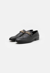 Černé kožené slip-on mokasíny s špičatými špičkami a zlatým řetízkem přes vrchní část, umístěné vedle sebe na bílém pozadí.