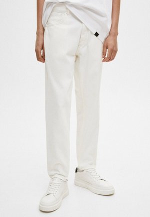 Osoba nosząca luźne, kremowe dżinsy, białe sneakersy z czarnymi akcentami na pięcie oraz częściowo wpuszczoną gładką białą koszulkę.