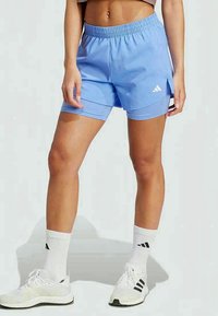 Kvinna som bär ljusblå Adidas två-i-ett sportshorts, vita strumpor och vita Adidas löparskor, stående mot en enkel bakgrund.