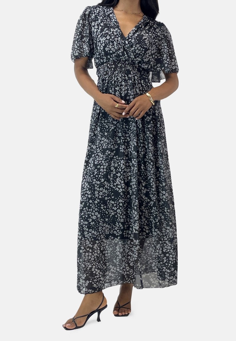 Femme portant une robe maxi noire et blanche à fleurs avec de courtes manches ballon et des sandales noires à talons avec des lanières.