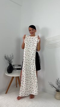 Frau, die ein langes weißes Kleid mit kleinen roten Blumendrucken hält, barfuß auf einem weißen Teppich in einem minimalistischen Raum steht.