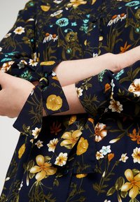 Blus med blommönster i marinblått tyg med gula, vita och gröna blommor. Har knytna ärmar, knapdetailjer och en lös passform.