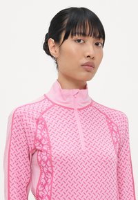 Lys rosa treningsgenser med halv glidelås og mønstret design i mørkere rosa, med en tettsittende silhuett og teksturerte detaljer langs sømmene.