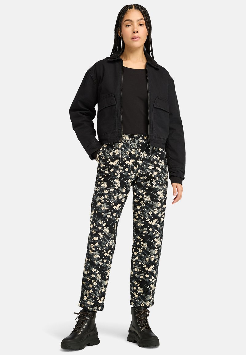 Giacca nera corta, top nero aderente, pantaloni floreali con fiori bianchi e verdi e stivali neri con lacci e suola spessa.