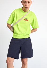 Neongrön t-shirt med framgrafik och korta ärmar, kombinerad med marinblå shorts och en ljusbeige axelväska.