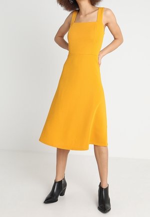 Femme portant une robe jaune sans manches, à encolure carrée, longueur genou avec jupe évasée et bottines noires, debout les mains sur les hanches.
