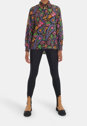 Person iført en langærmet, farverig blomster-paisley skjorte med sorte leggings og tan og sorte ankelstøvler, der står mod en hvid baggrund.