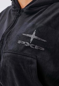 Sudadera con cremallera negra de tela suave, con un diseño de estrella adornado con rhinestones en el lado izquierdo del pecho y un bolsillo frontal.
