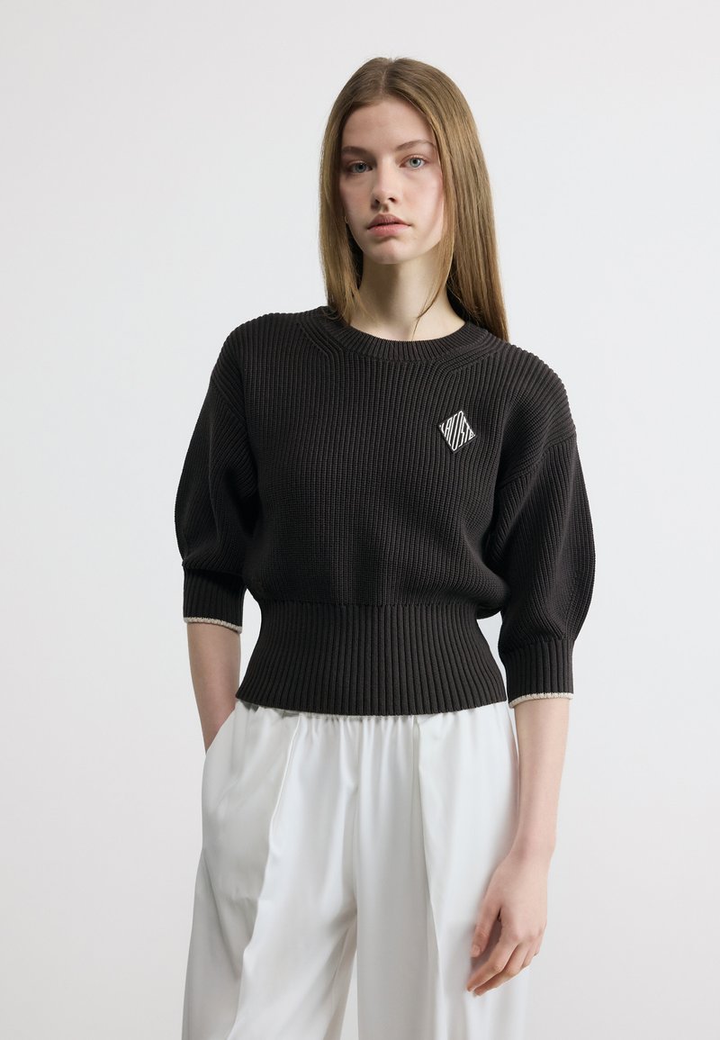 Νεαρή γυναίκα με ίσια μαλλιά φοράει μαύρο ριπ πλεκτό με λογότυπο στο στήθος και φαρδιά λευκά παντελόνια, στέκεται μπροστά σε ανοιχτό φόντο.