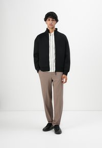 Joven se presenta de pie, mirando hacia adelante, vistiendo una chaqueta negra con cremallera, camisa blanca con rayas verticales, pantalones beige y zapatos negros.