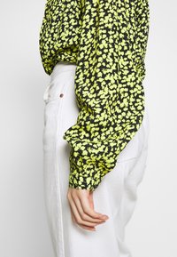 Květnová halenka v černé barvě s jasně žlutými vzory, volného střihu, s nabíranými rukávy a manžetami, spojená s bílými kalhotami.