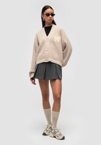 Beżowy sweter robiony na drutach z over-size'owymi rękawami, pięcioma guzikami i naszywką w kształcie litery 'W'. Połączony z szarą spódnicą plisowaną i kremowymi skarpetkami do kostek.