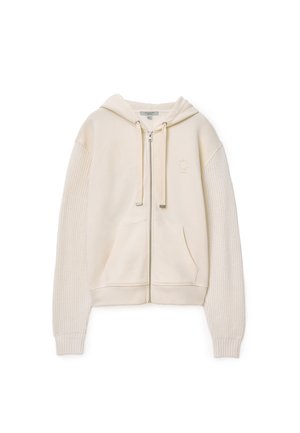 Felpa con zip color crema, dotata di cappuccio e maniche con texture a maglia. Materiale del corpo liscio con tasche frontali e dettaglio logo sottile.
