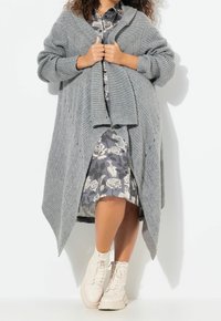 Světlý šedý pletený oversized kardigan s žebrovanou texturou, otevřeným předním dílem a šálovým límcem, nošený přes květinové šaty a béžové šněrovací boty.