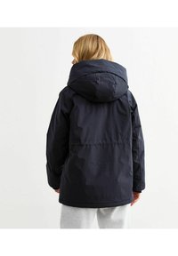 Navyblauwe gewatteerde jas met een grote capuchon, elastische manchetten en een ontspannen pasvorm, ontworpen voor warmte en comfort.