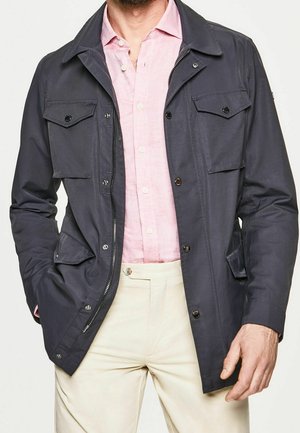 Mężczyzna w rozpiętej ciemnoszarej kurtce na jasnoróżowej koszuli zapinanej na guziki i beżowych spodniach, z rękami w kieszeniach kurtki.