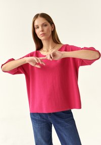 Maglione oversize fucsia con scollo ampio, maniche a tre quarti e tessuto strutturato, abbinato a jeans blu.