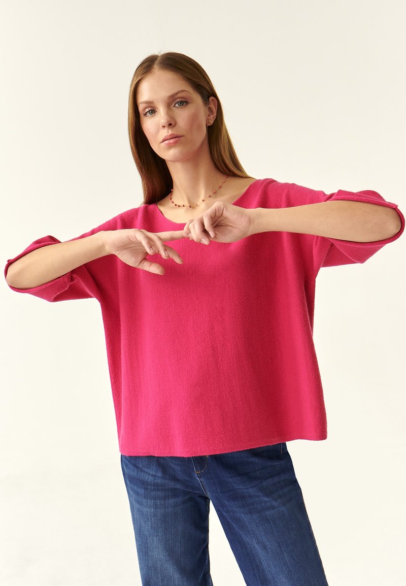 Maglione oversize fucsia con scollo ampio, maniche a tre quarti e tessuto strutturato, abbinato a jeans blu.
