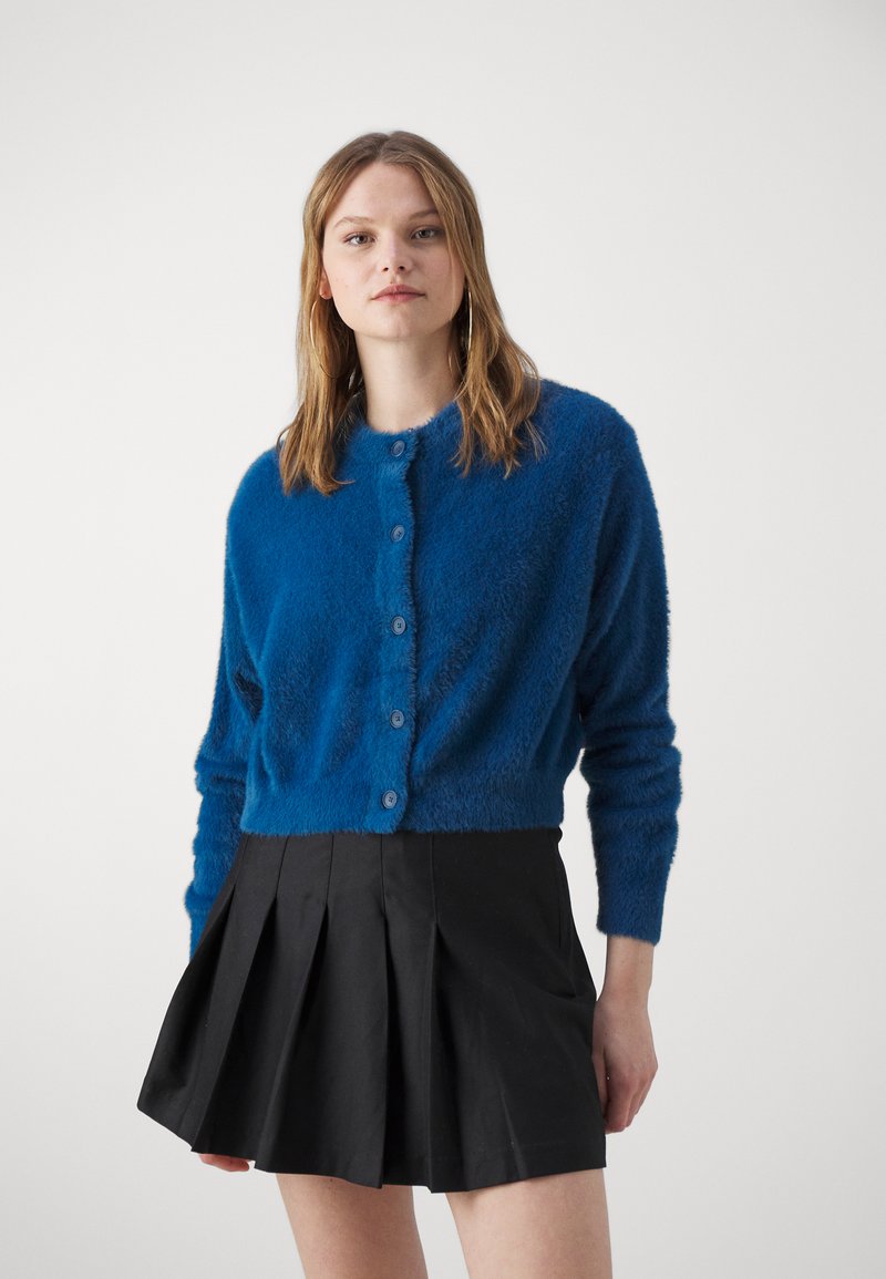 Cardigan blu fluffy con scollo rotondo e chiusura a bottoni. Indossato sopra una gonna nera plissettata, evidenziando un contrasto di texture e colore.