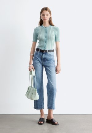 Nainen pukeutunut mintunvihreään raidalliseen lyhythihaiseen napilliseen toppiin, korkeavyötäröisiin sinisiin farkkuihin, mustiin slip-on-sandaaleihin ja pitää vaaleanvihreää käsilaukkua.