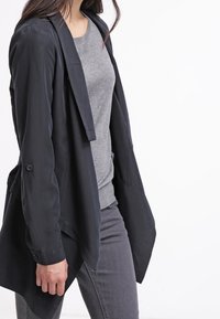 Černý blazer s dlouhým rukávem a volným střihem, vyrobený z hladké látky, v kombinaci se šedým svetrem a přiléhavými šedými kalhotami.