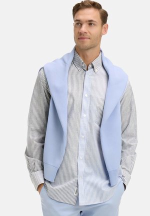 Homme portant une chemise à rayures bleu clair et blanches avec un pull bleu clair posé sur les épaules, les mains dans les poches.