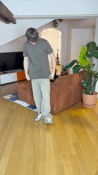 Camisa polo cinza, calças cargo verde-claro e ténis prateados; de pé num chão de madeira numa sala de estar com plantas e móveis.